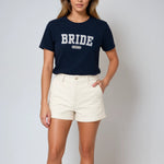 Woman wearing a navy 'BRIDE' t-shirt with white shorts on a plain background