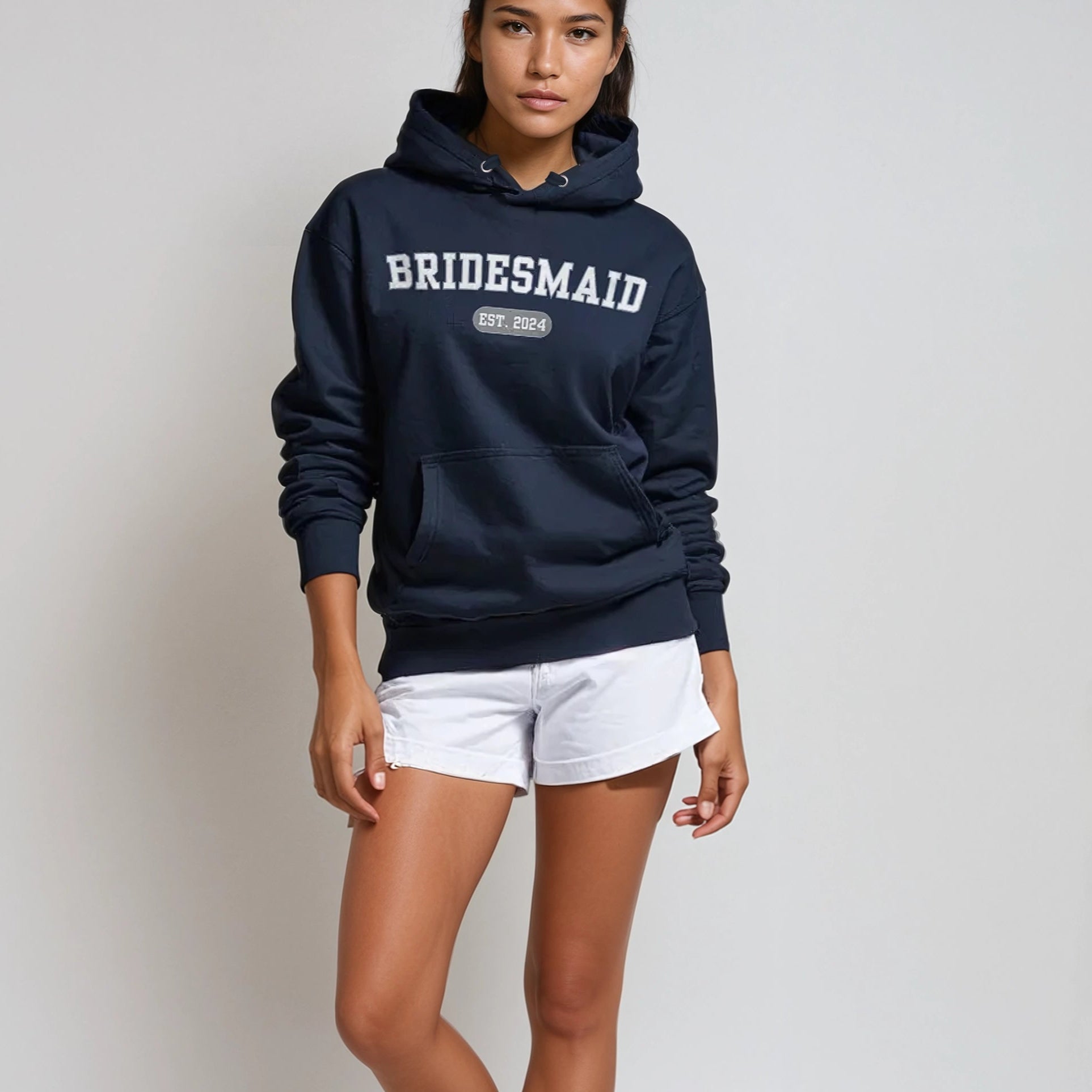 Woman wearing a navy hoodie with 'BRIDESMAID' text, white shorts, and white sneakers on a plain background