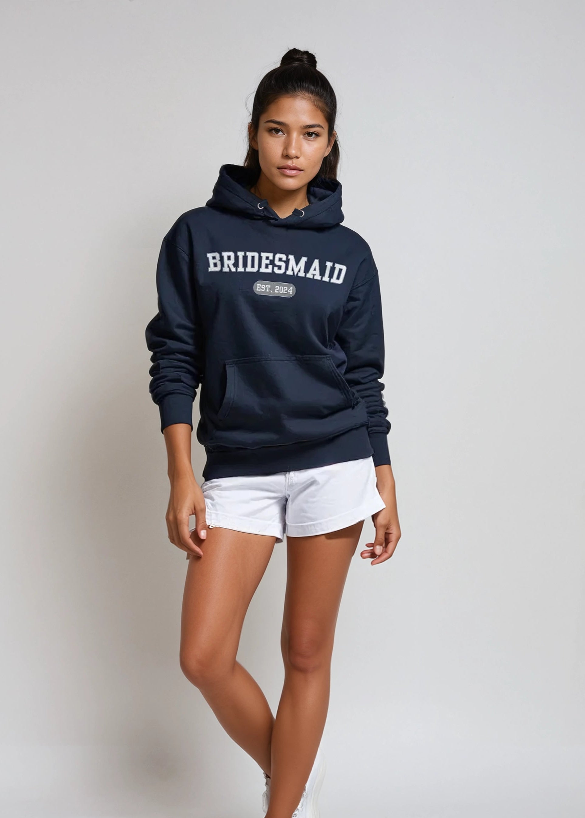 Woman wearing a navy hoodie with 'BRIDESMAID' text, white shorts, and white sneakers on a plain background