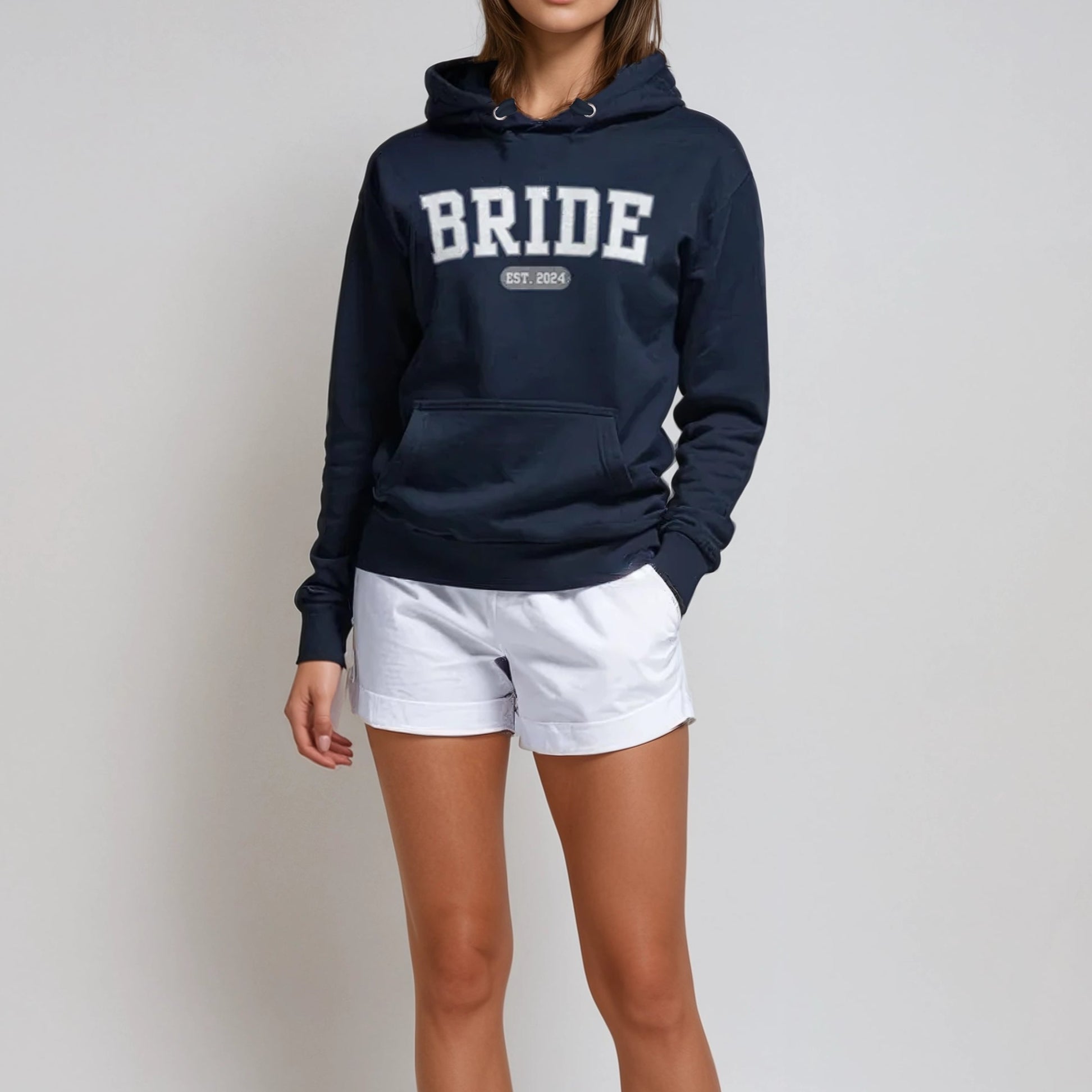 Woman wearing a navy hoodie with 'BRIDE' text, white shorts, and white sneakers on a light gray background