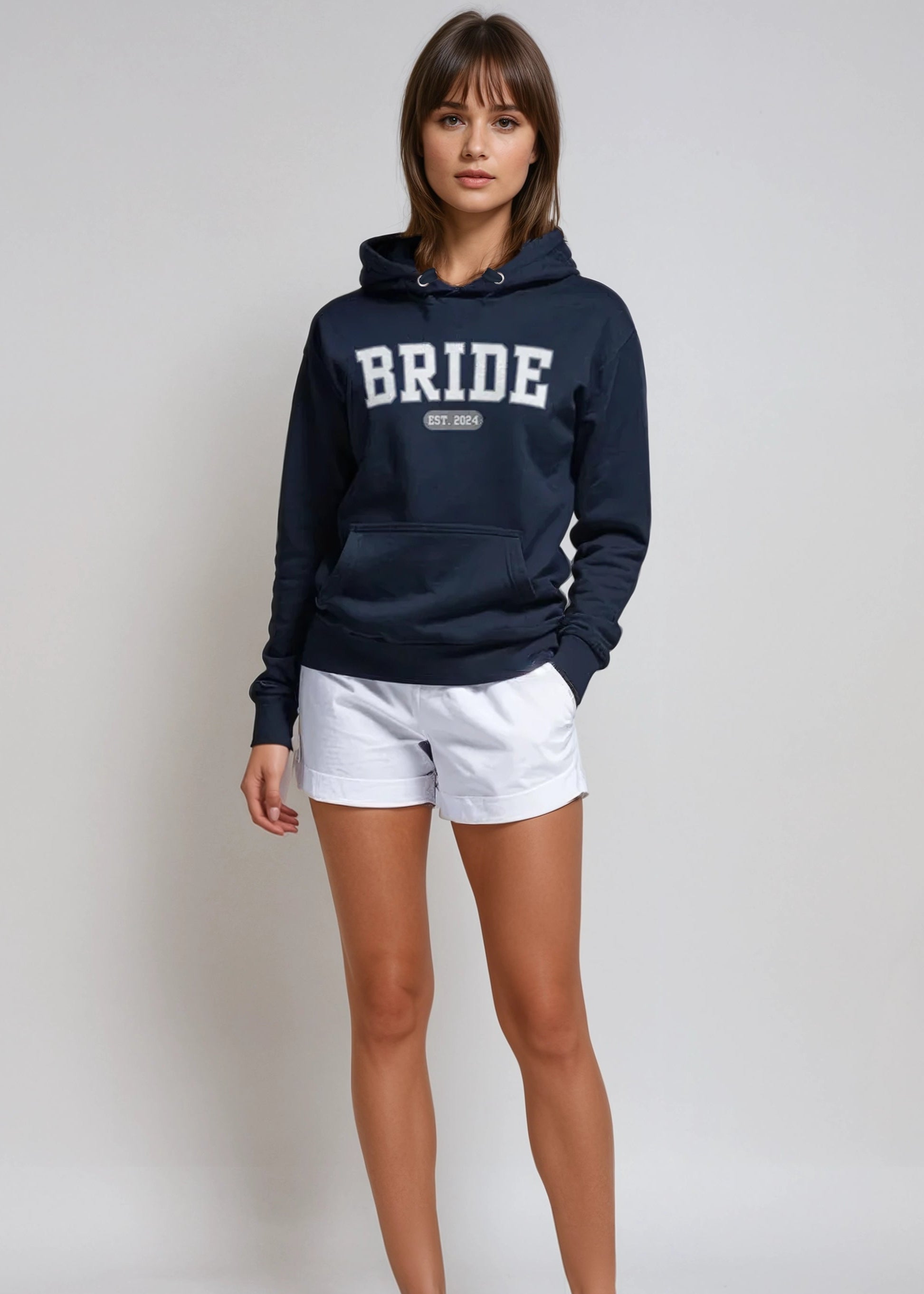 Woman wearing a navy hoodie with 'BRIDE' text, white shorts, and white sneakers on a light gray background