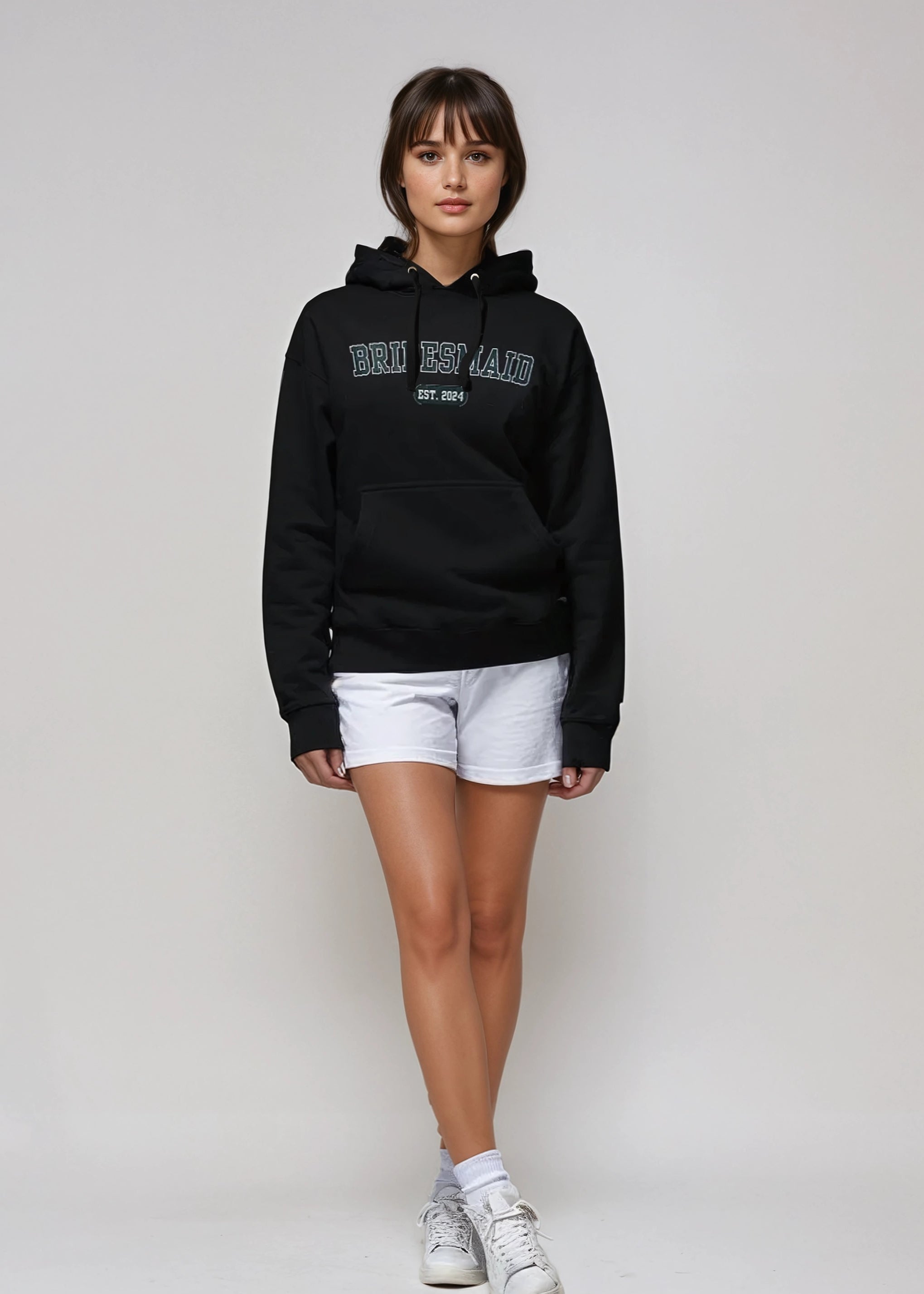 Person wearing a black hoodie with text, white shorts, and white sneakers on a plain background